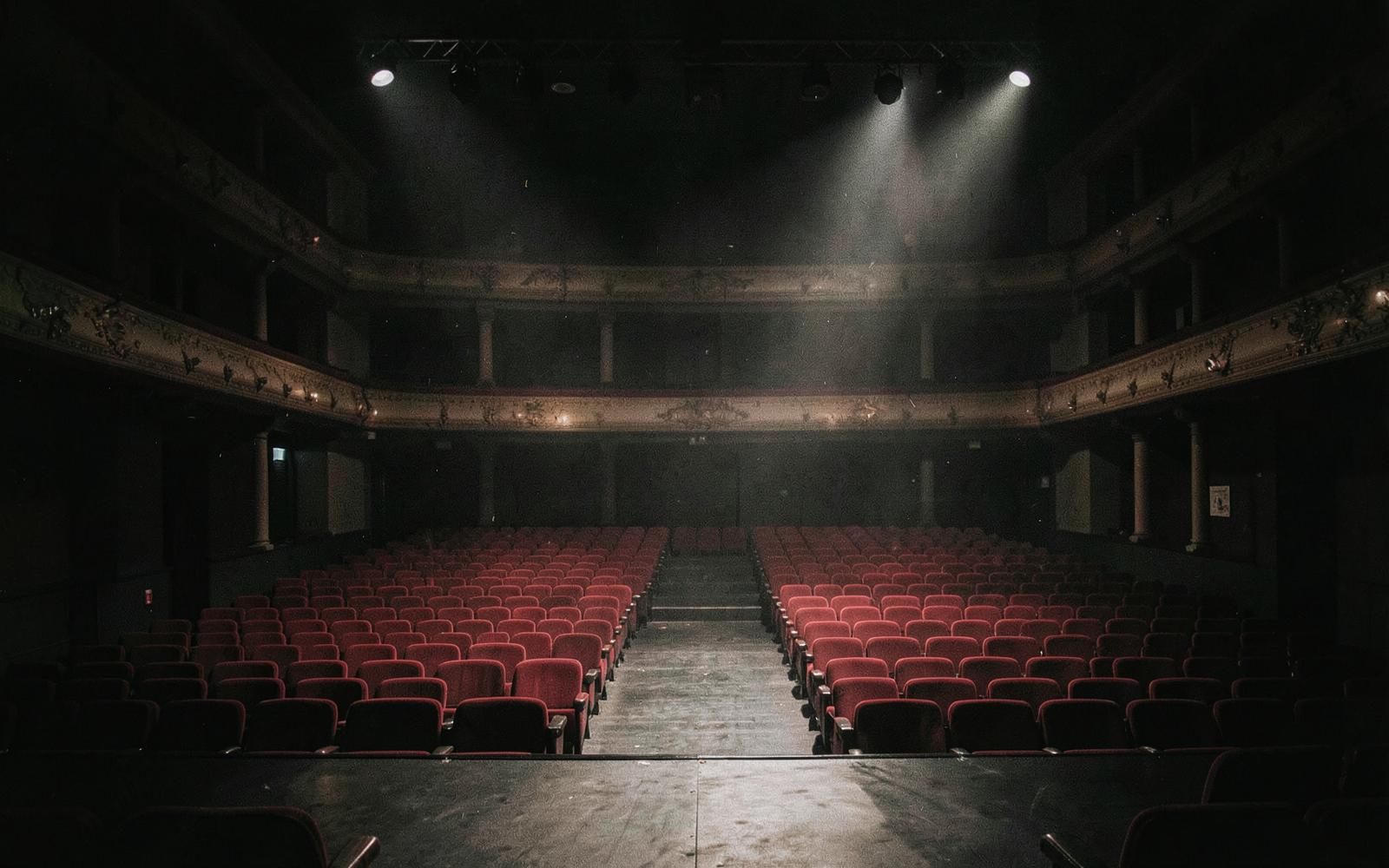Empty theatre auditorium in low light, seen from the stage, red seats muted by shadow